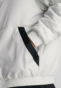 Veste blanche matelassée avec un accent de poche latérale noire. Dispose de poignets élastiques et d'un ourlet froncé pour un ajustement confortable. Texture douce et lisse.