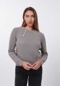 Grå ribbestrikket genser med lange ermer, rund halslinning og dekorative knapper på skulderen. Brukes over svarte bukser.