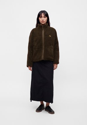 Femme debout portant une veste polaire zippée marron foncé, une longue jupe noire et des baskets noires devant un fond clair uni.