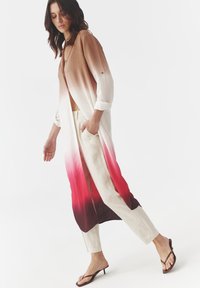 Dlouhý gradientový kimono v odstínech hnědé, krémové a červené, vyrobený z lehkého materiálu, s ohrnutými rukávy a uvolněným střihem. Spárovaný s béžovými kalhotami.