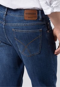 Jeans de mezclilla azul oscuro con una etiqueta de cintura de cuero marrón. Notable costura en los bolsillos traseros en un patrón en forma de V.