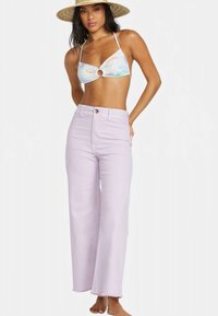 Woman wearing wide-brimmed hat, pastel floral bikini top with center ring, and high-waisted light lavender wide-leg jeans, standing barefoot.