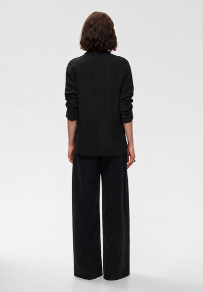 Personne aux cheveux courts et bruns, vue de dos, portant une chemise noire à manches longues avec les manches retroussées et un pantalon noir large sur un fond uni.
