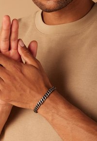 Zwarte en zilveren gevlochten armband met een zachte textuur, gedragen om een pols. De achtergrond toont een beige stof met een gestructureerde uitstraling.