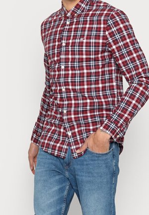 Mężczyzna w koszuli na guziki w czerwono-biało-niebieską kratę z długim rękawem, w niebieskich dżinsach, z ręką w kieszeni, stojący na jednolitym tle.