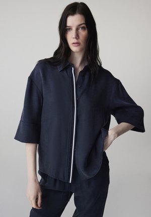 Vrouw met lang donker haar, gekleed in een losse marineblauwe blouse met witte bies en bijpassende broek, staand met één hand op haar heup tegen een effen achtergrond.