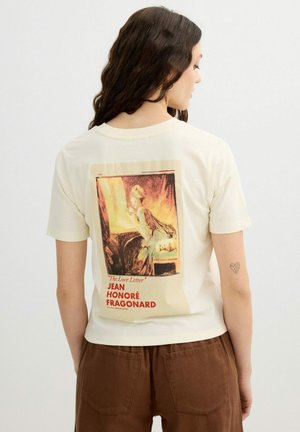 Mujer con cabello largo y oscuro, que lleva una camiseta crema con una pintura de estilo vintage titulada "La carta de amor" de Jean Honoré Fragonard en la espalda.