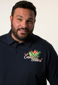 Námořnická polo košile vyrobená z bavlny s texturovaným povrchem. Obsahuje vyšívaný design "Exotický ostrov" s tropickými listy a oranžovými květy.