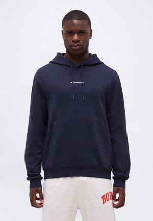 Homme portant un sweat à capuche bleu marine avec poche avant et un short blanc avec un texte rouge, debout devant un fond clair uni.