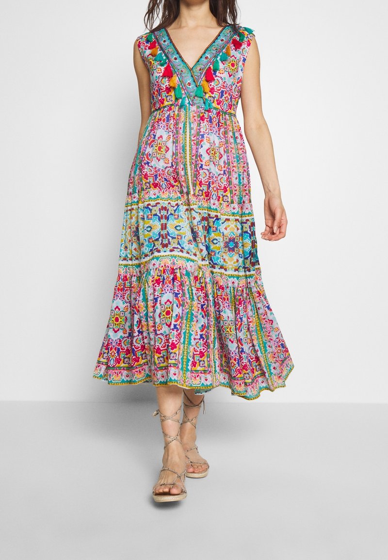 Abito maxi floreale multicolore con scollo a V, maniche a cappuccio e orlo a strati. Presenta motivi intricati e nappine colorate sullo scollo.