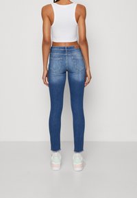 Mujer vistiendo jeans skinny azul de cintura alta, top corto blanco, calcetines blancos y zapatillas blancas con toques en pastel, de pie contra un fondo blanco.