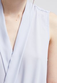 Top senza maniche azzurro chiaro con design avvolgente, realizzato in tessuto morbido. Presenta un collo a V e una sottile lucentezza. Nessun motivo o accessorio visibile.