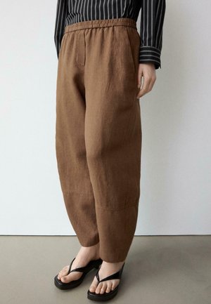 Osoba ubrana w luźne brązowe spodnie z elastyczną talią, czarną bluzkę w paski z długim rękawem oraz czarne klapki na platformie, stojąca w pomieszczeniu.