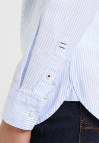 Jasnoniebieska i biała w paski koszula z długim rękawem, z zapięciem na guzik przy mankiecie, z małym czerwono-niebieskim logo na rękawie oraz zaokrąglonym dołem.