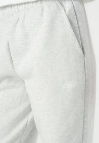 Pilkos medvilninės sportinės kelnės su elastinga juosmeniu ir kišenėmis, ant kairės pusės yra iškilus New Balance logotipas. Minkšta tekstūra.