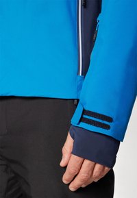 Helle blaue Outdoor-Jacke aus strukturiertem Stoff, mit verstellbaren Bündchen mit Klettverschluss und einer Reißverschlusstasche an der Seite.