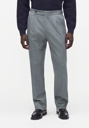 Homme portant un pantalon gris taille haute avec des boutons d'ajustement latéraux, associé à des mocassins en cuir noirs et un haut sombre à manches longues.