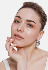 Orecchini in argento a forma organica con finitura lucida. Modella che tiene il mento, con trucco naturale e acconciatura elegante.