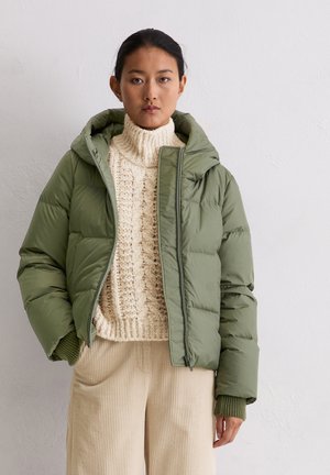 Olivgrüne Steppjacke mit Kapuze, ausgestattet mit einem vorderen Reißverschluss und einer gesteppte Textur. Getragen über einem cremefarbenen Zopfpullover und beigen Cordhose.