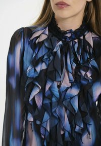 Zila un melna ruffle blūze ar augstu apkakli un slāņveida auduma tekstūru. Smalka gradienta raksts ar izteiktu ruffle detalizāciju.