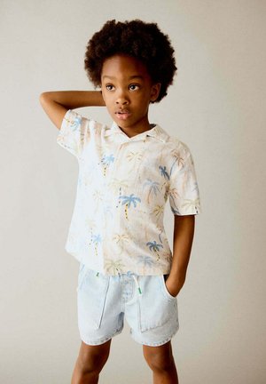 Jeune garçon aux cheveux bouclés portant une chemise à manches courtes palmier et un short en jean clair, debout avec une main derrière la tête.