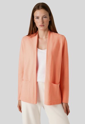 Junge Frau mit glatten braunen Haaren, die eine korallenfarbene offene Strickjacke über einem weißen Top und weißen Hosen trägt und vor einem grauen Hintergrund steht.