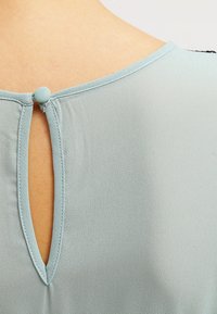 Encolure dos d'un chemisier bleu clair avec un petit bouton recouvert de tissu et une ouverture en forme de goutte.