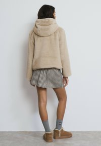 Chaqueta de piel sintética beige con capucha, combinada con una minifalda de cuadros. Botines marrones con detalle de ribete y calcetines grises completan el conjunto.