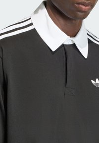 Čierna polokošeľa s bielym golierom a tromi bielymi pruhmi na ramenách. Na ľavej strane hrudníka je vyšívané logo. Hladká textúra.