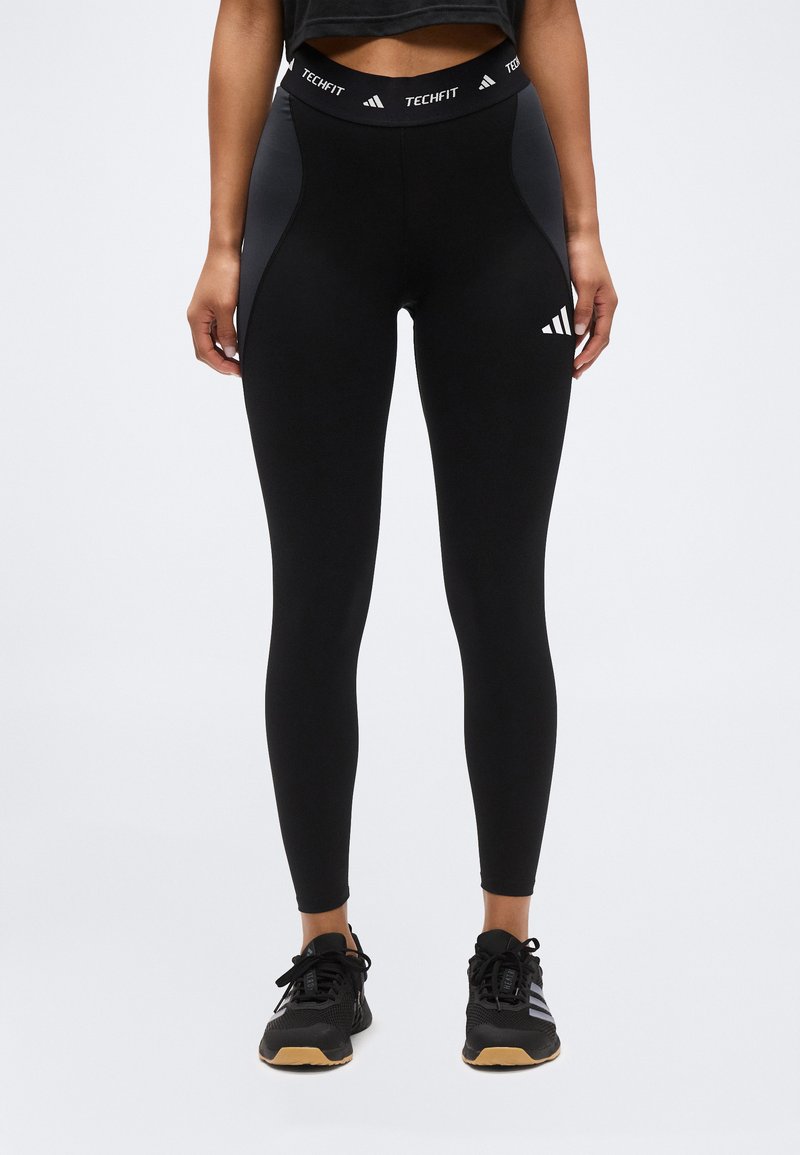 Γυναίκα που φοράει μαύρα κολάν Adidas Techfit και μαύρα αθλητικά παπούτσια, στέκεται με τα χέρια χαλαρά στα πλευρά μπροστά σε απλό φόντο.