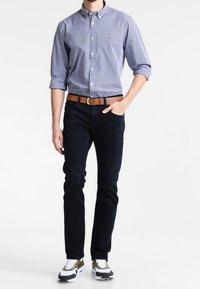 Camisa de cuadros vichy azul marino, jeans oscuros, cinturón marrón y zapatillas blancas con detalles en oliva. Diseño ajustado, mangas remangadas y cuello clásico.