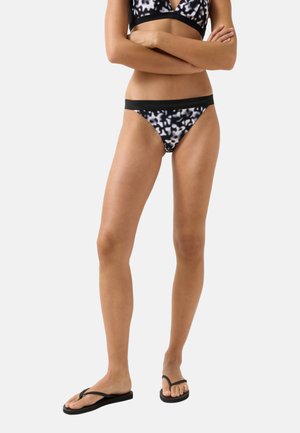 Kobieta ubrana w czarno-białe wzorzyste spodnie i górę od bikini z czarną lamówką, stoi ze skrzyżowanymi ramionami, nosi czarne klapki.
