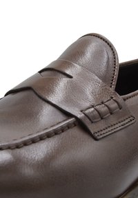 Braune Lederloafers mit gesticktem Detail und einem dekorativen Riemen über dem Spann. Glatte Textur mit glänzendem Finish.