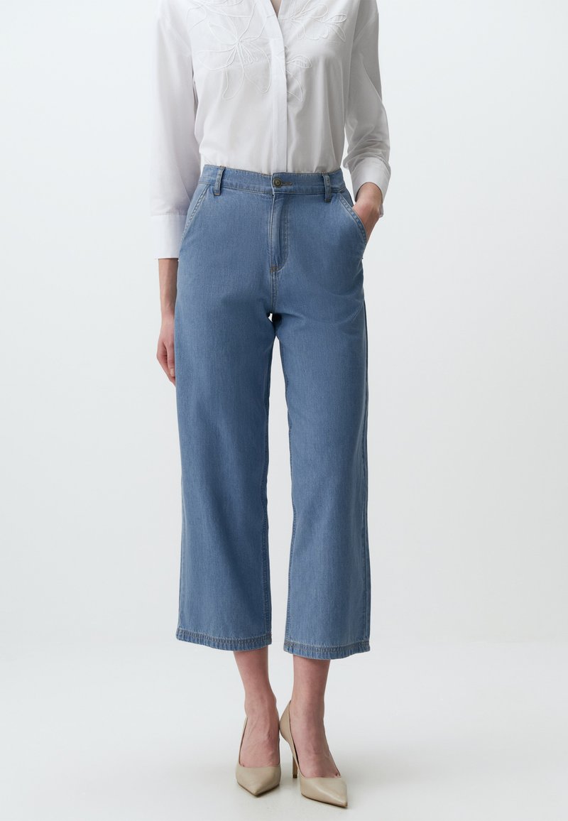 Jean wide-leg en denim bleu clair avec taille haute, comprenant des poches avant et une fermeture à bouton, associé à un chemisier blanc brodé.