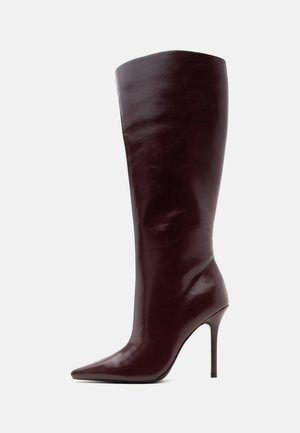 Botte en cuir marron foncé montant jusqu’au genou avec bout pointu et talon haut fin sur fond clair.