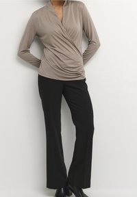 Langärmliges, Wickeloberteil aus beigem Stoff mit drapierter Vorderseite. Kombiniert mit hochgeschnittenen schwarzen Hosen und Stiefeletten, die eine schlanke Silhouette kreieren.