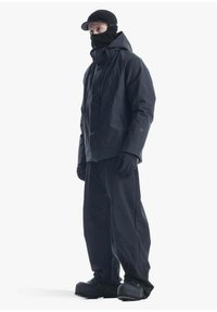 Completo di abbigliamento esterno nero composto da una giacca con cappuccio e zip frontale, pantaloni ampi, guanti e calzature grandi con suole testurizzate.