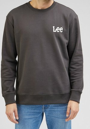 Homme portant un sweat-shirt Lee gris foncé et un jean bleu, debout avec les bras relâchés le long du corps.