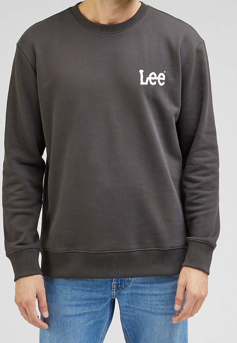 Homme portant un sweat-shirt Lee gris foncé et un jean bleu, debout avec les bras relâchés le long du corps.