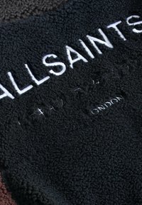 Juodas vilnos audinys su iškilmingomis baltomis raidėmis "ALLSAINTS" ir įspaustomis detalėmis "LONDINAS", demonstruojantis tekstūruotą paviršių ir spalvų kontrastą.