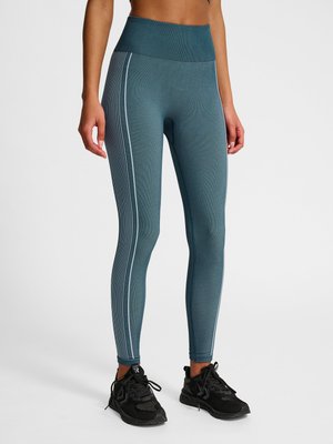 Vrouw draagt hooggetailleerde blauw-grijze legging met verticale witte strepen aan de zijkanten en zwarte sportschoenen, staand op een witte achtergrond.