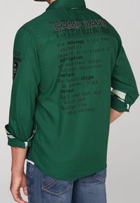 Chemise verte à coupe décontractée, avec un texte imprimé dans le dos et des accents à motifs sur les manches et le col. Matière en mélange de coton.