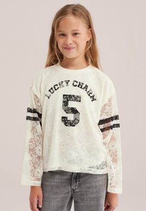 Une fille porte un haut blanc en dentelle à manches longues, orné de rayures noires et d’un grand chiffre 5, assorti à un jean gris.