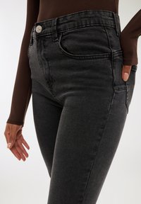 Zbliżenie na osobę w czarnych dżinsach z wysokim stanem i brązowej bluzce z długim rękawem, z jedną ręką blisko kieszeni, a drugą ręką w luźnej pozycji.