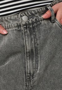Gros plan de mains dans les poches sur un jean en denim noir délavé taille haute avec bouton argenté, porté avec un haut rayé noir et blanc.