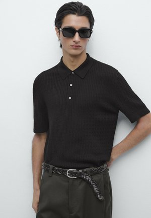 Young man wearing black textured short-sleeve polo, dark braided belt, dark pants, and black rectangular sunglasses against a plain white background.