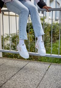 Białe sneakersy na platformie z różowymi akcentami, teksturowana skóra, sznurowany design i wyraźne logo na języku, siedzące na poręczy.