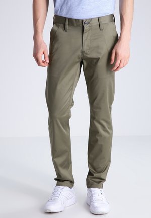 Mann trägt schlank geschnittene olivgrüne Hose und weiße Turnschuhe, steht vor einem schlichten hellen Hintergrund.