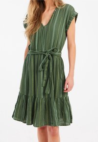 Grünes gemustertes Kleid mit V-Ausschnitt, kurzen Ärmeln und einem Bindegürtel. Das Kleid hat senkrechte Streifen mit Punkten und einen rüschenbesetzten Saum.