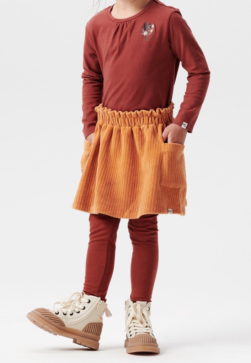 Camiseta de manga larga color óxido con un diseño floral, combinada con una falda de pana naranja con una cintura con volantes y bolsillos. Botas crema con cordones.